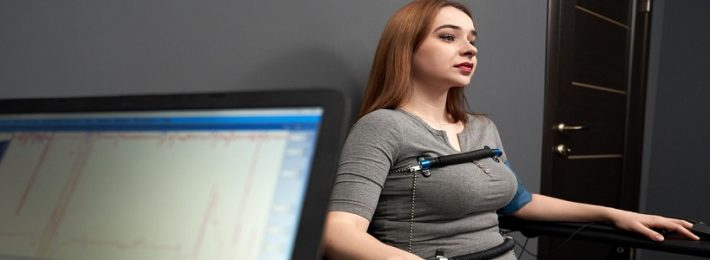 Young beautiful woman sitting with attached sensors on fingers, hands and body, testing with computer polygraph, looking away. Graph and polygraph on screen of lie detector